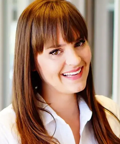A woman with long straight brown hair and bangs is smiling. She is wearing a white collared shirt and is standing in front of a blurred indoor background.